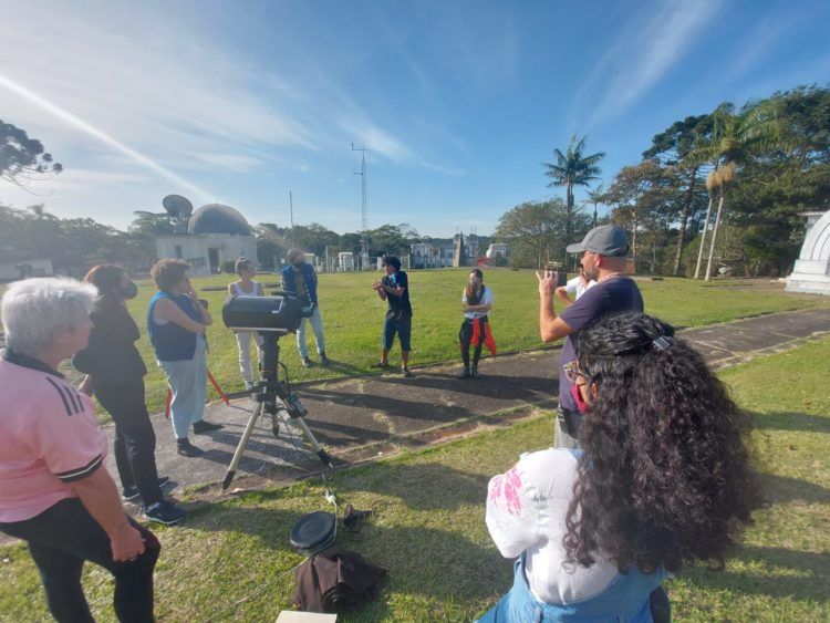 Grupo de pessoas reunidas em semicírculo, em área aberta no Parque CienTec, observando orientações de monitor sobre o uso do telescópio.