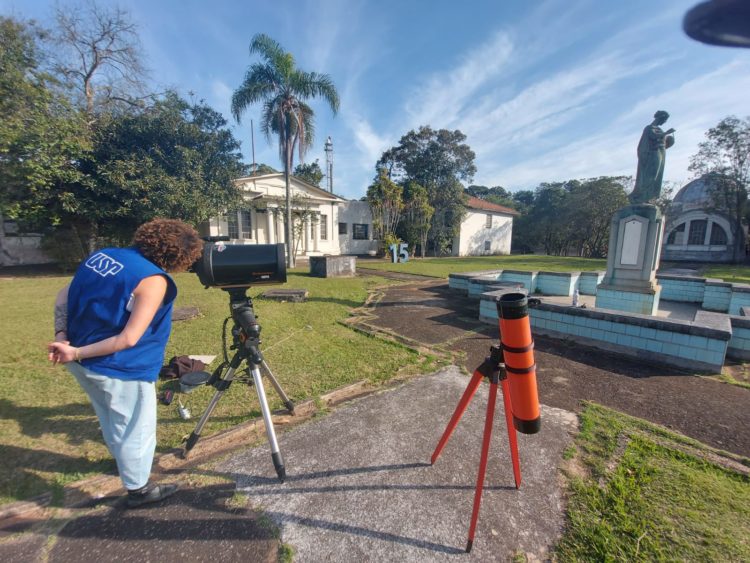 Monitora do Parque CienTec observa telescópio no canto esquerdo. Ao centro, outro telescópio, de cor laranja, com estátua da Urânia em segundo plano, no canto direito. 